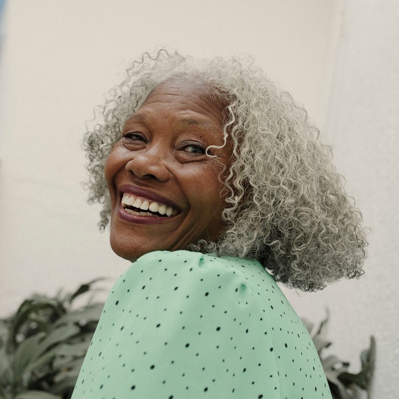 A smiling older woman with grey curly hair, photographed outdoors